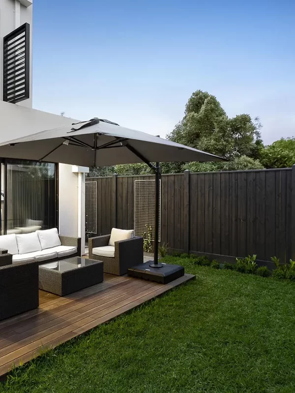 Modern two-story house with large sliding glass doors leading to an outdoor patio with a seating area and umbrella, surrounded by a well-manicured lawn and wooden privacy fence.