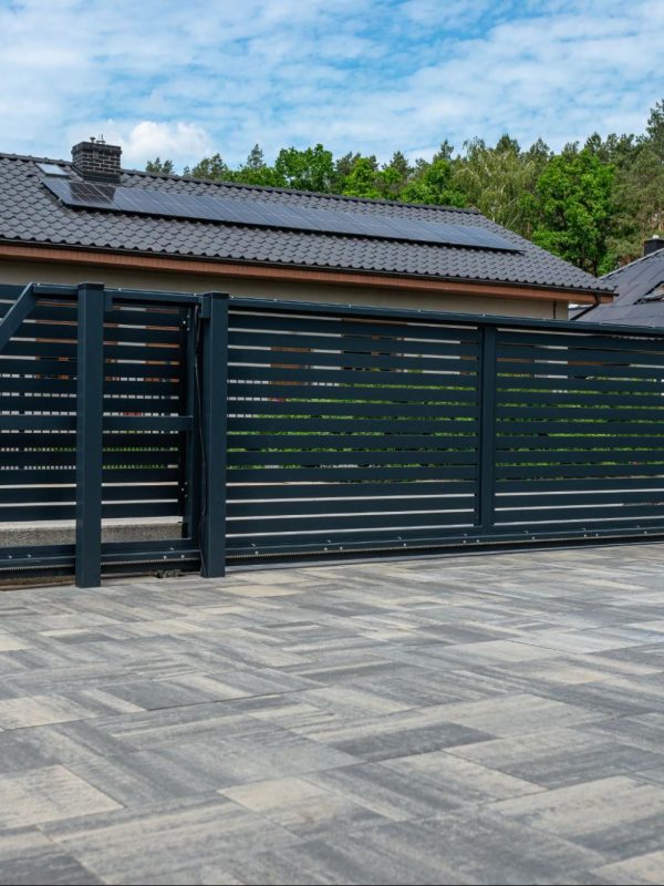 A modern anthracite-colored panel fence with a visible sliding gate leading to the garage and a slab driveway, with rows of houses visible behind it.