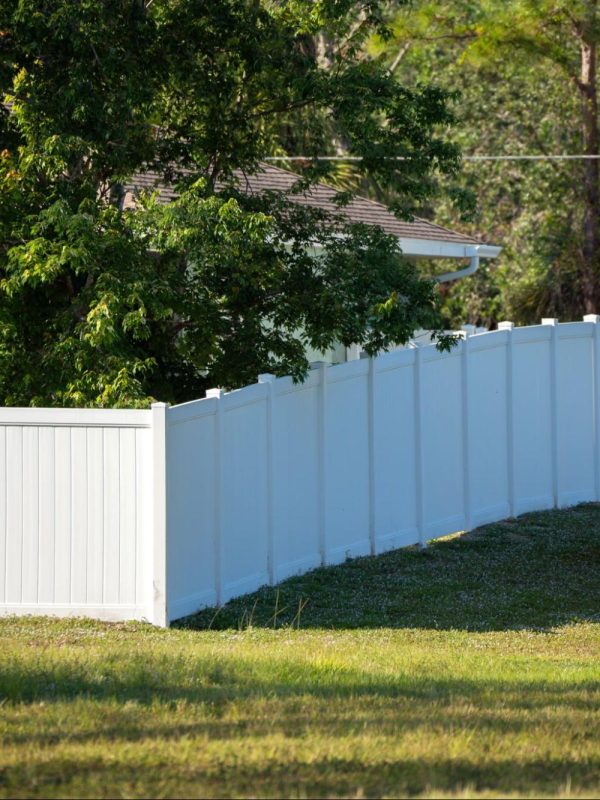 A white vinyl picket backyard fence encloses a vibrant green lawn, surrounded by tall, lush trees that create a tranquil and secure outdoor setting.