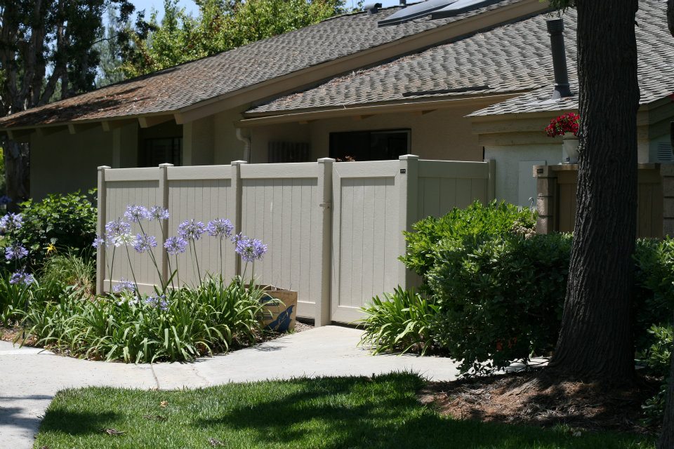 166060_430618240311643_304441850_n A beige fence in front of a house with purple flowers and greenery growing alongside it.
