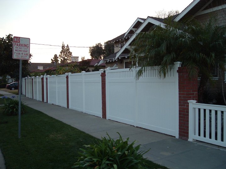 27759_116664778373659_3629163_n A residential street with a white fence and a 'No Parking' sign on the left side, and houses with palm trees on the right side.