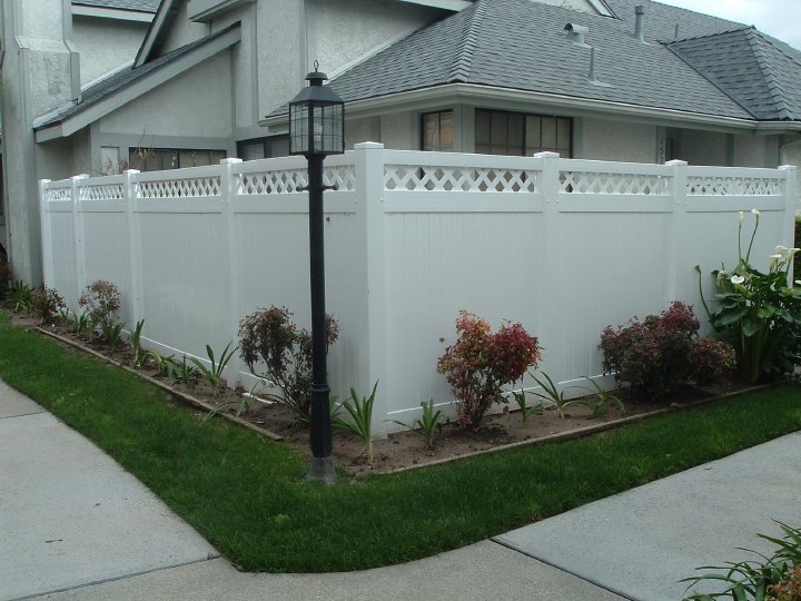 image 1 A white vinyl privacy fence with a decorative lattice top along the perimeter of a house's front yard, with a line of small shrubs planted in front and a black lamp post on the left side.