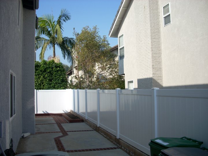 image 2 A narrow pathway between two beige houses with a white fence on one side and palm trees in the background.
