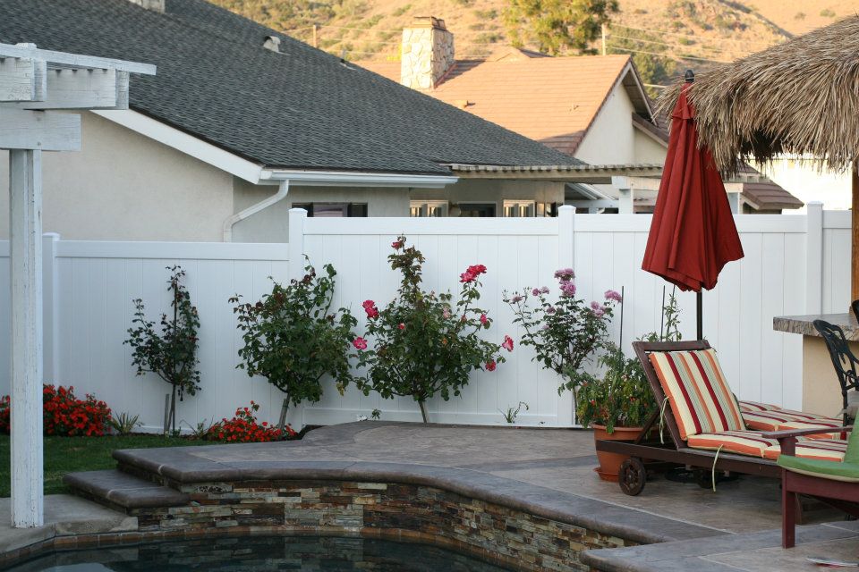 image 3 A well-manicured backyard with a white fence, blooming rose bushes, a pool with a stone edge, and a lounging chair with red and green stripes next to a closed red umbrella.