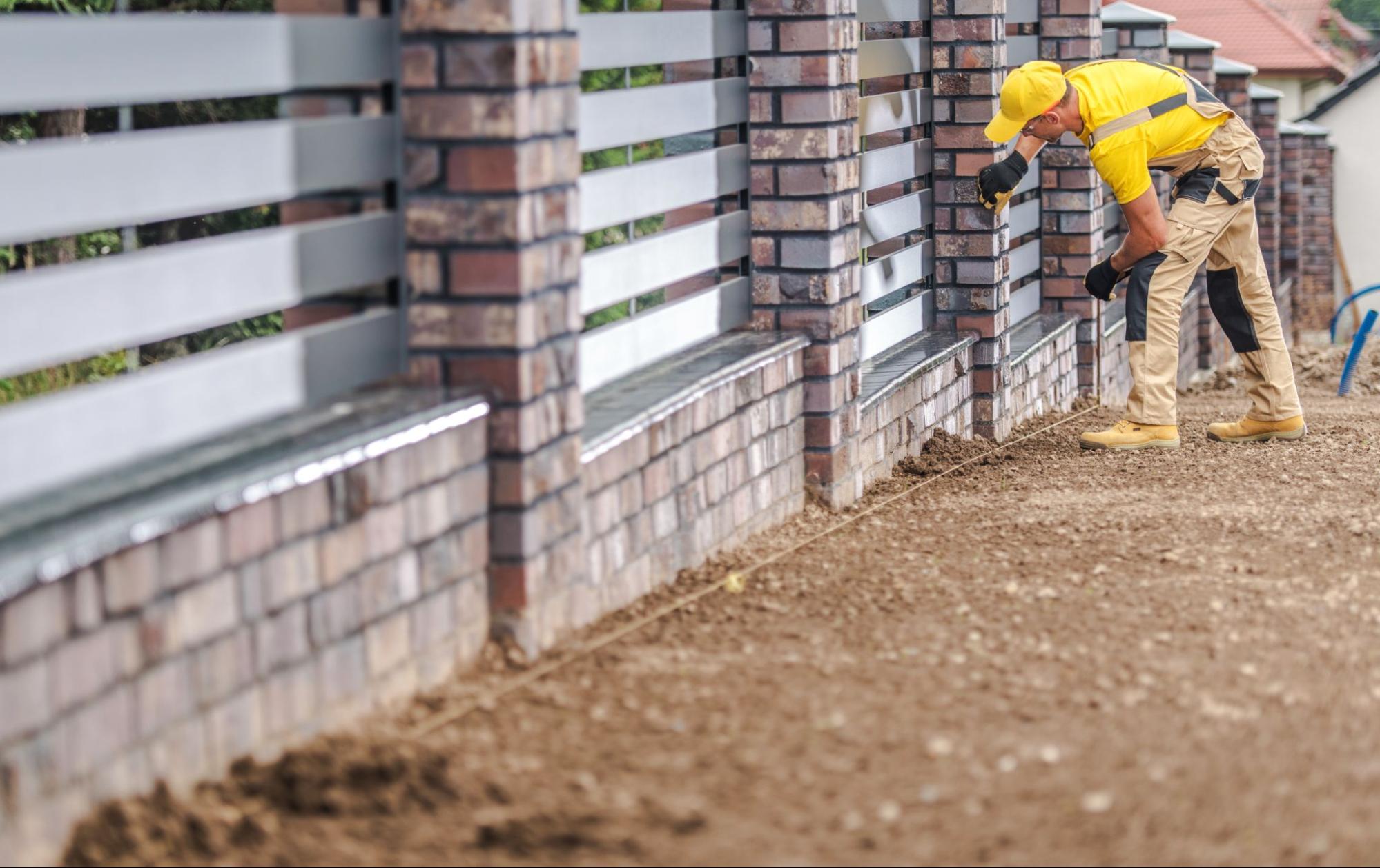 A male worker in cargo overalls, with a yellow shirt underneath and a cap, is seen assembling a decorative residential fence.