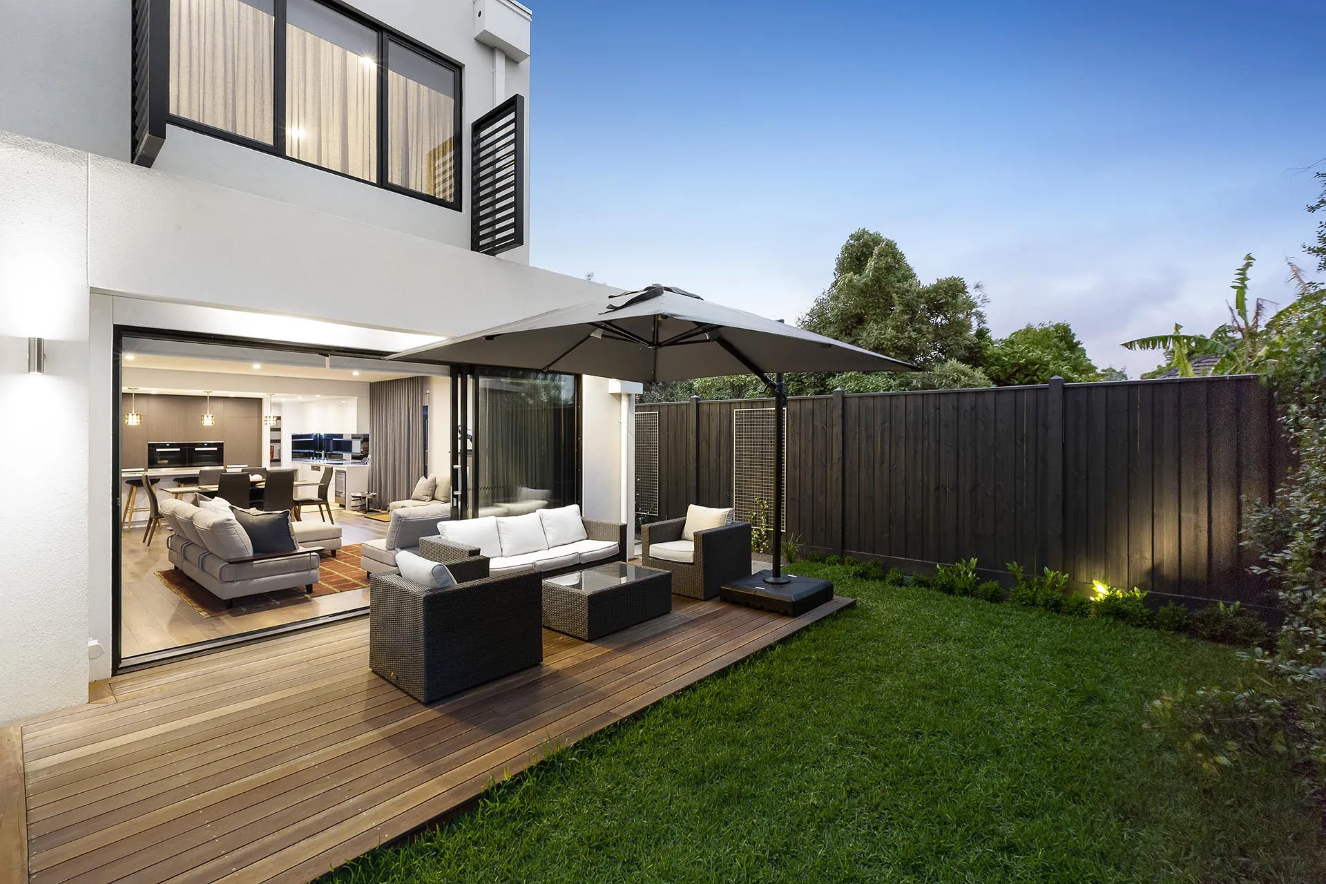 Modern two-story house with large sliding glass doors leading to an outdoor patio with a seating area and umbrella, surrounded by a well-manicured lawn and wooden privacy fence.