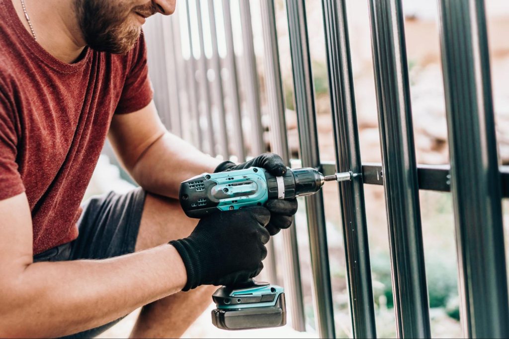 A man uses a cordless drill to fasten a screw into a metal fence while wearing gloves.